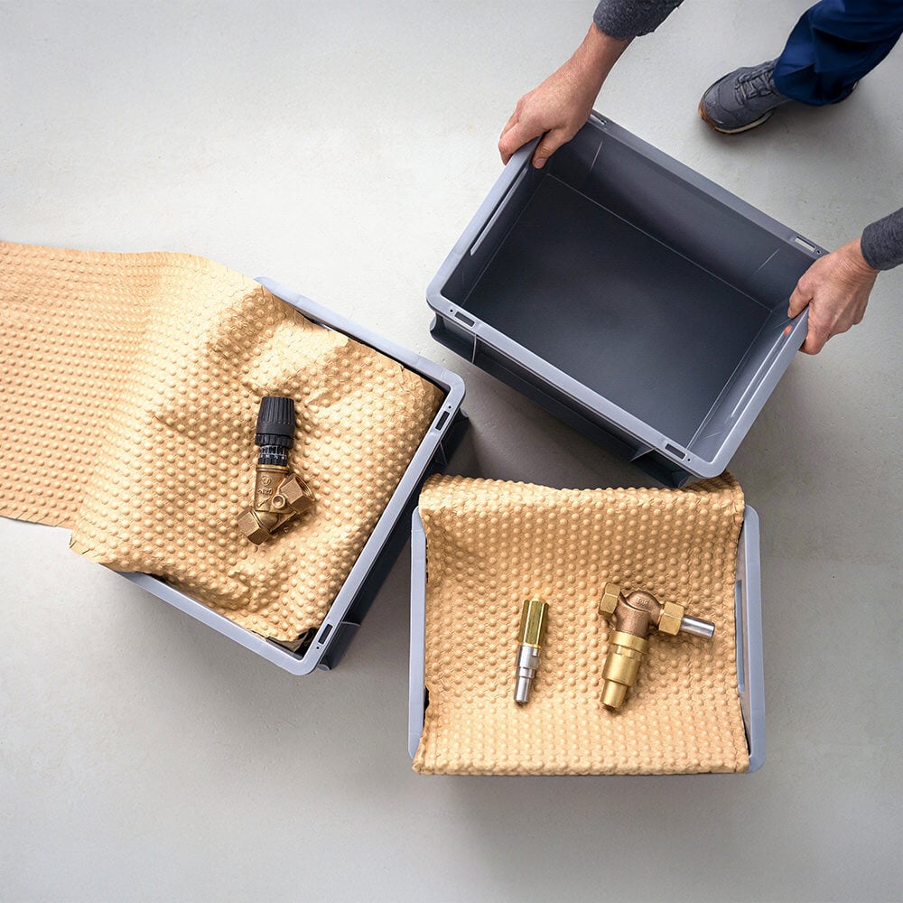 Ein Produktionsarbeitender stellt einen Kleinlastträger zu zwei weiteren mit in PapairWrap gepolsterten technischen Gegenständen.