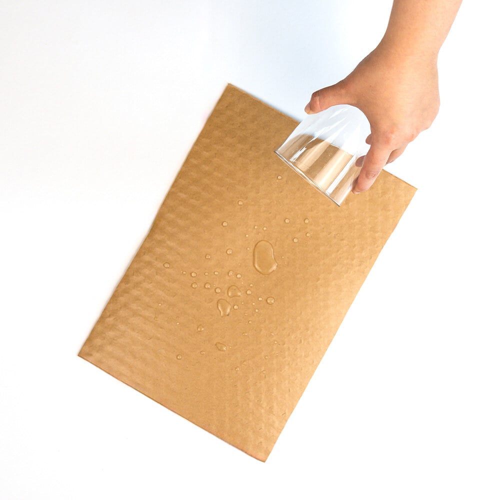Eine Hand schüttet Wasser aus einem Glas auf einen Bogen beschichtetes PapairWrap Plus und die Tropfen perlen ab.
